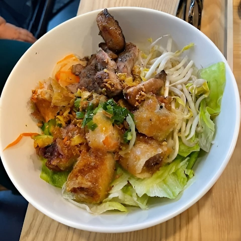 A white bowl of Vietnamese vermicelli with grilled pork, crispy spring rolls, and fresh vegetables.