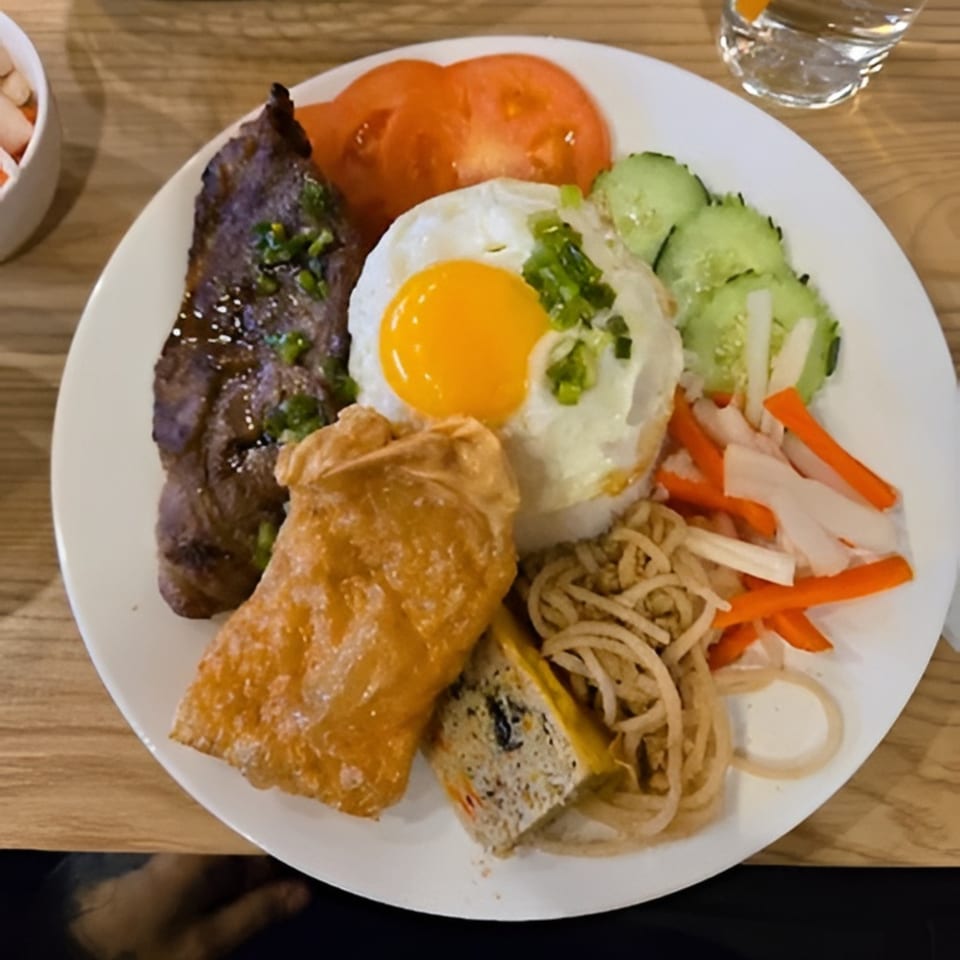 A Vietnamese grilled pork chop with rice, fried egg, egg roll, and pickled vegetables on a white plate.