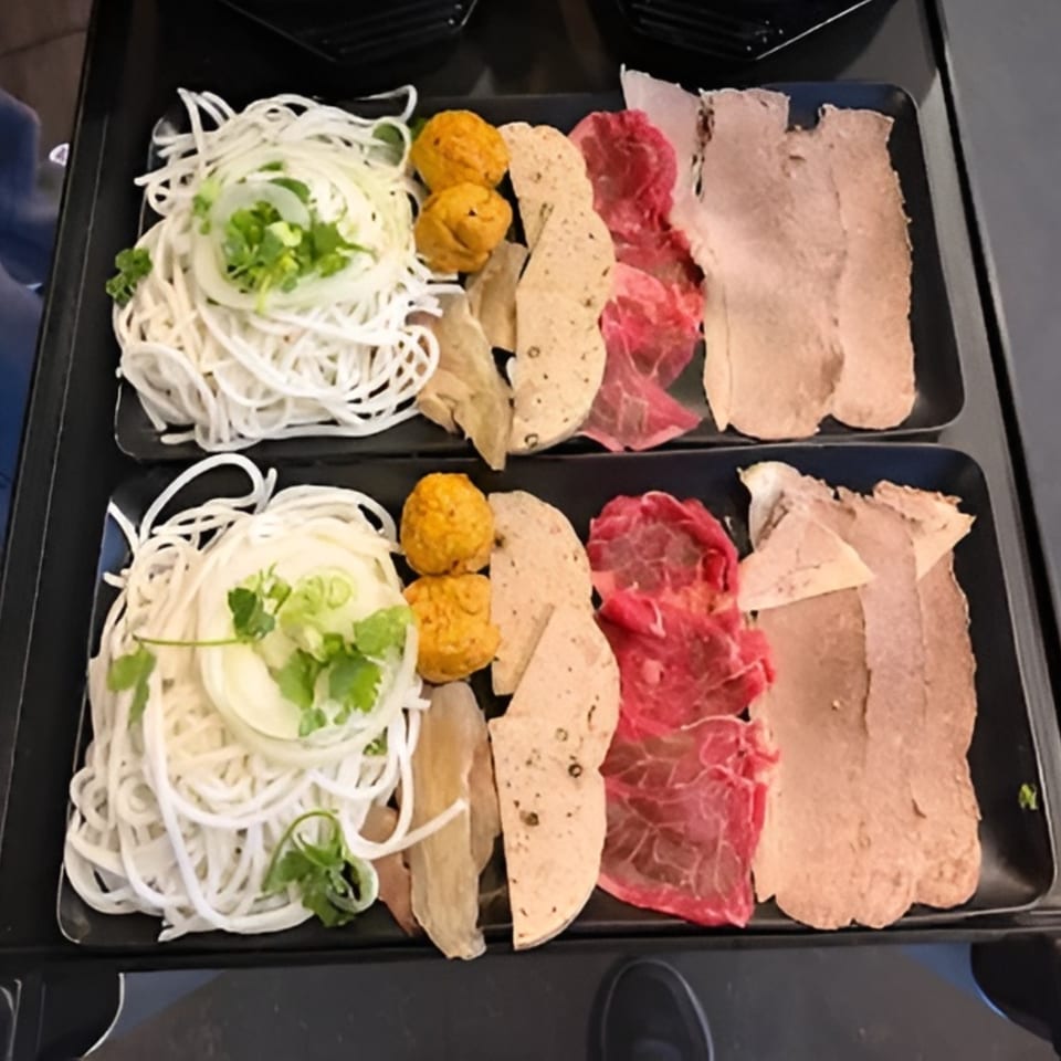 Two black trays filled with pho ingredients: noodles, onions, cilantro, meatballs, sliced meats, and beef.